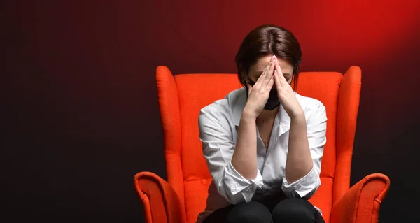 A stressed woman holds her head while sitting in a red chair, during a test and a coronavirus pandemic. The concept of viral danger and fear