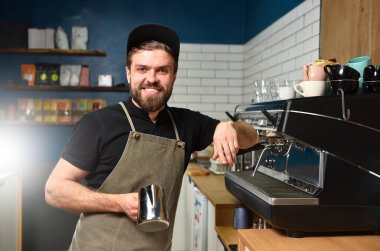 Genç sakallı bir barmen kahve makinesinin yanındaki kafede gülümsüyor..
