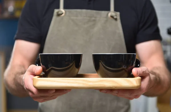 Barista bartender guy in a coffee shop holds two mugs of coffee in his hands