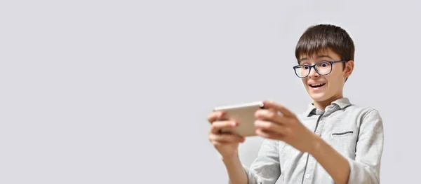 Teenager mit Brille auf grauem Hintergrund spielt auf dem Smartphone ein Spiel mit Überraschungsmomenten. — Stockfoto