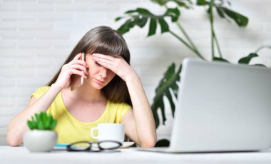 Dizüstü bilgisayarda çalışan yorgun bir kız ev ofisinde telefonla konuşuyor. Uzak çalışma konsepti