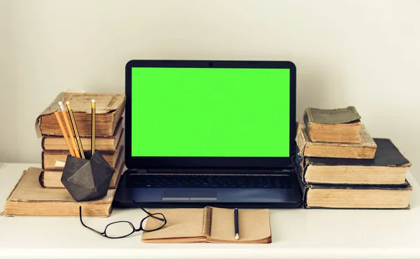 Green screen laptop, stack of old books, notebook and pencils on