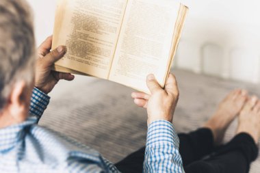 Orta yaşlı, gri gözlüklü, mavi gömlekli, kitap okuyan bir adam. 