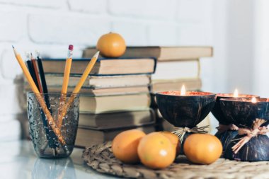 Hindistan cevizi kabuğunda spa aromalı mumlar, hasır tezgahta mandalinalar, cam beyaz bir masada plastik tutacakta kitap ve kalemler, eğitim kavramında samimi ev içi arka plan.