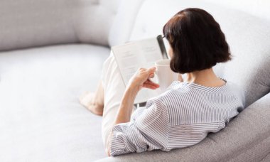 Orta yaşlı, esmer, gözlüklü, gri koltukta kitap okuyan kadın.