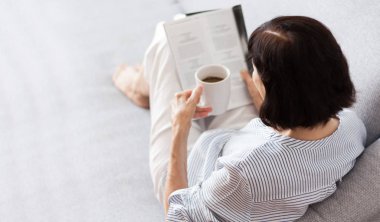 Orta yaşlı, esmer, gözlüklü, gri koltukta kitap okuyan kadın.