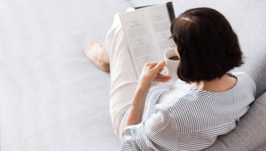 Orta yaşlı, esmer, gözlüklü, gri koltukta kitap okuyan kadın.