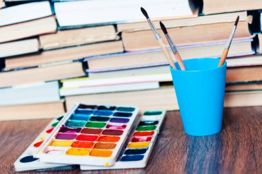 Suluboya boya, iyi kullanılmış fırçalar ve arka planda kitap yığını olan ahşap masa üzerindeki kağıt.