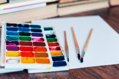 Suluboya boya, iyi kullanılmış fırçalar ve arka planda kitap yığını olan ahşap masa üzerindeki kağıt.