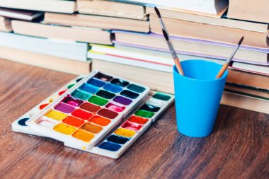 Suluboya boya, iyi kullanılmış fırçalar ve arka planda kitap yığını olan ahşap masa üzerindeki kağıt.