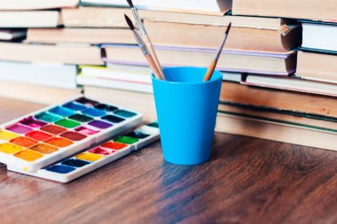 Suluboya boya, iyi kullanılmış fırçalar ve arka planda kitap yığını olan ahşap masa üzerindeki kağıt.