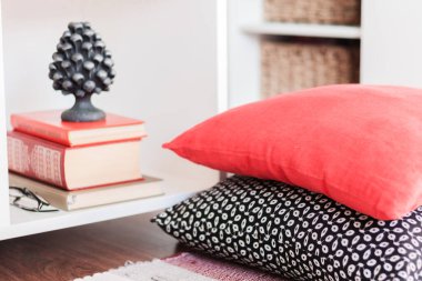 Çok renkli bir halı ve dekoratif yastıkların yanında mum ve kitaplarla dolu beyaz bir komodin.