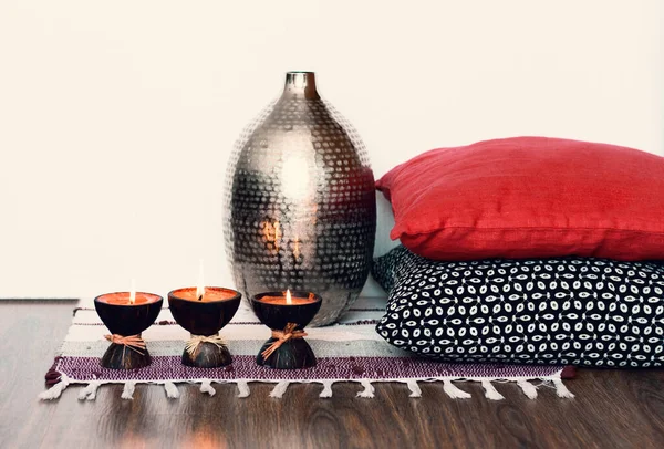Cozy home interior decor, burning spa aroma candles in coconut shell on a multi-colored rug, near metal vase and decorative pillows