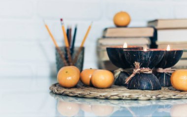 Hindistan cevizi kabuğunda spa aromalı mumlar, hasır tezgahta mandalinalar, cam beyaz bir masada plastik tutacakta kitap ve kalemler, eğitim kavramında samimi ev içi arka plan.