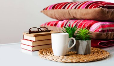 Konforlu ev dekorasyonu: bir fincan kahve, kitap yığını, hasır tezgahtaki saksılarda bitkiler, yastıklar ve beyaz masadaki ekoseler. Uzaktan eğitim. Karantina konsepti:
