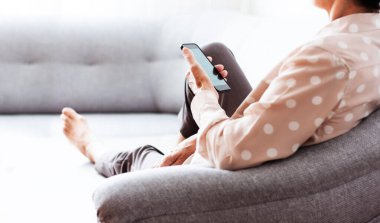 Gri koltukta oturan orta yaşlı esmer kadın cep telefonu akıllı telefonu kullanıyor, internette sörf yapıyor.