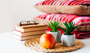 Konforlu ev dekorasyonu: Elma, kitap yığını, hasır tezgahtaki saksılar, yastıklar ve beyaz masadaki ekoseler. Uzaktan eğitim. Karantina konsepti: