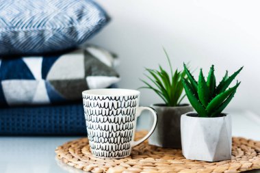 Konforlu ev dekorasyonu: beyaz ve siyah bardak ve odadaki beyaz bir masanın sehpasında duran tencerelerdeki süs bitkileri. Arka planda mavi yastıklar. Karantina konsepti: