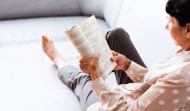 Orta yaşlı esmer kadın gri kanepede kitap okuyor, yumuşak odaklı, ev kavramında kalıyor, rahat bir geçmişi var.