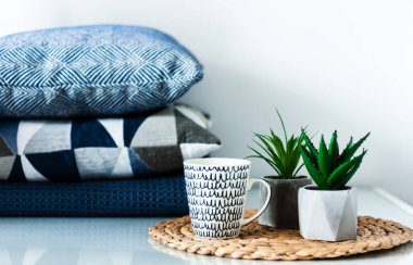 Konforlu ev dekorasyonu: beyaz ve siyah bardak ve odadaki beyaz bir masanın sehpasında duran tencerelerdeki süs bitkileri. Arka planda mavi yastıklar. Karantina konsepti: