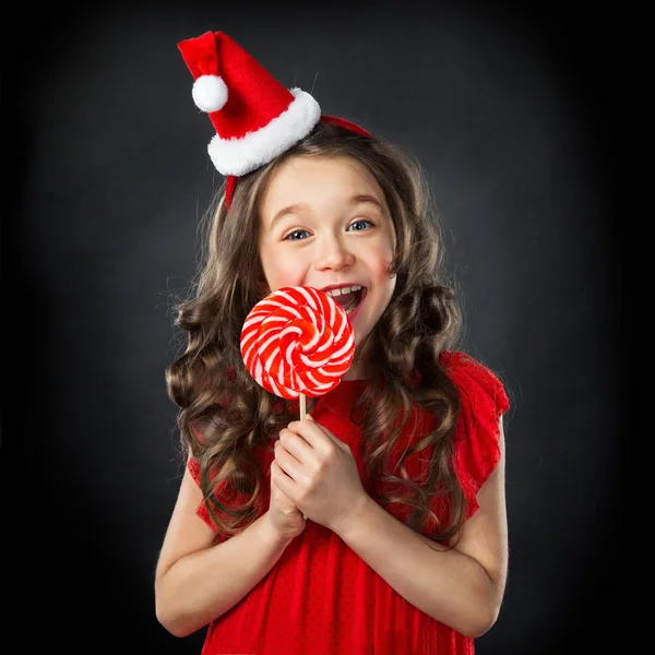 Niña sonriente en sombrero de santas con caramelo, fondo oscuro aislado —  Fotos de Stock