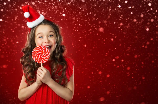 Klein meisje in kerstmuts met snoep op rode achtergrond. Kersttijd — Stockfoto
