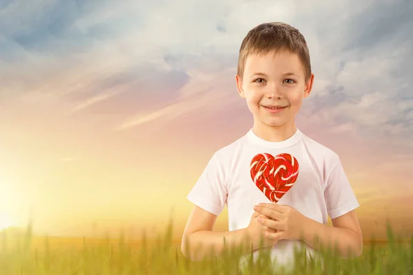 Menino com pirulito vermelho doce em forma de coração no fundo da natureza — Fotografia de Stock