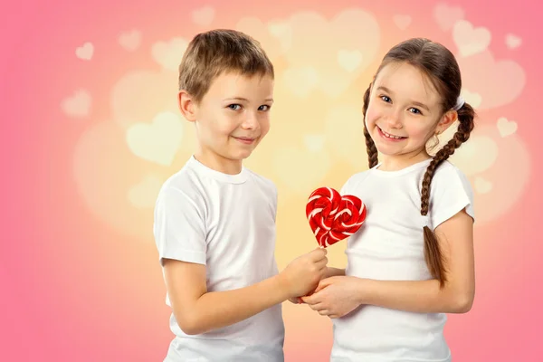 Niños, niño le da a una niña caramelo lollipop rojo en forma de corazón sobre fondo rosa —  Fotos de Stock
