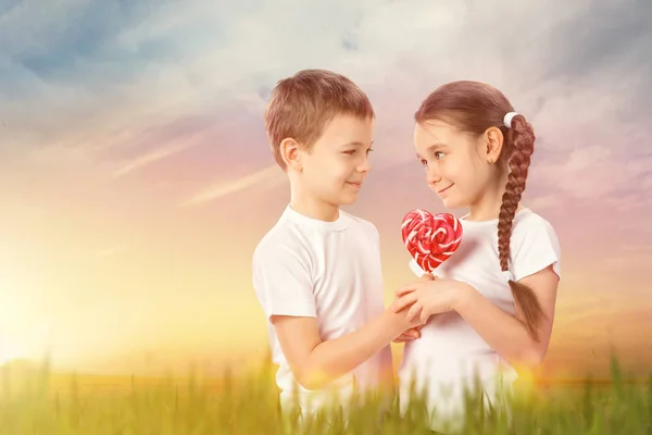 Boy le da a una niña caramelo lollipop rojo en forma de corazón en el campo de atardecer. Día de San Valentín —  Fotos de Stock
