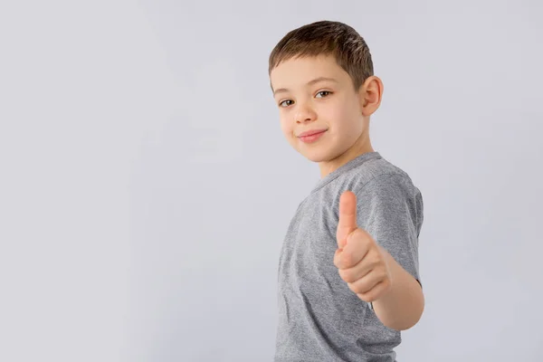 Mały chłopiec pokazuje kciuk gest w szary T-shirt na białym tle. — Zdjęcie stockowe