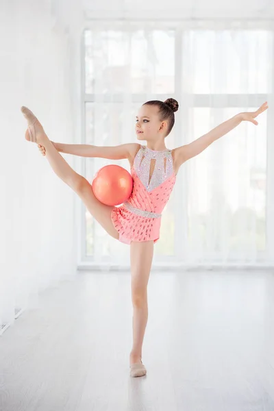 Красива маленька дівчинка-гімнастка в рожевому спортивному одязі, виконуючи елемент художньої гімнастики з м'ячем у фітнес-класі — стокове фото