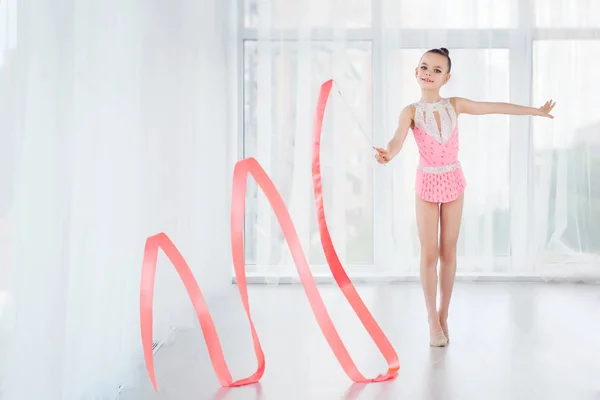Beautiful little gymnast girl in pink sportswear dress, doing rhythmic gymnastics exercise Spirals with art ribbon