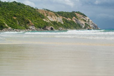 Aşağıdan, beyaz kumlu bir kumsal ve yeşil bir tepenin arka planına karşı gelgitten gelen küçük bir dalga..