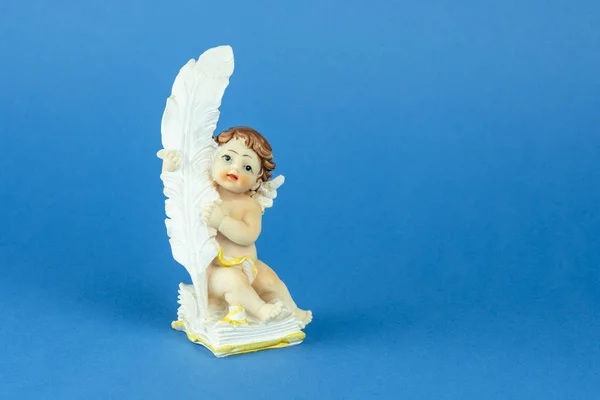 Figurine of a little sitting angel with a large feather in his hands on a blue background.