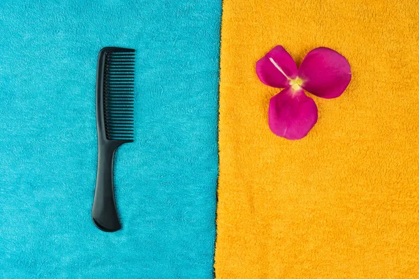 A black hairbrush lies on a blue towel and next to it is a pink rose petal on a yellow towel.