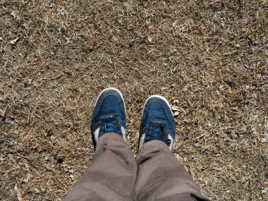 Kahverengi pantolonlu erkek bacakları ve yerde kuru otlu mavi spor ayakkabılar..