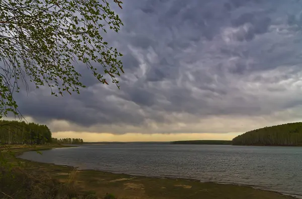 Lake Baikal.Spring.The sky is covered with thick gloomy volumetric clouds.The water receded from the shore of the lake, and the bottom was exposed.On the shore the hummocks are covered with thick grass