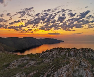 Doğu Sibirya. Baykal Gölü. Aya Körfezi. Pembe, turuncu, sarı sayısız kümülüs bulutu zümrüt ve mavi gökyüzünde yükselen güneş tarafından aydınlatılıyor. Güneşin ışıkları. Sudaki yansımalar.