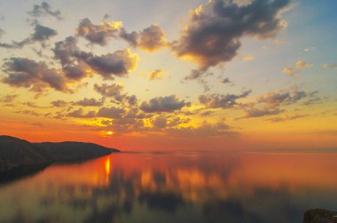 Doğu Sibirya. Baykal Gölü. Aya Körfezi. Pembe, turuncu, sarı sayısız kümülüs bulutu zümrüt ve mavi gökyüzünde yükselen güneş tarafından aydınlatılıyor. Güneşin ışıkları. Sudaki yansımalar.