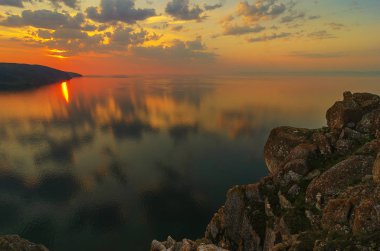 Doğu Sibirya. Baykal Gölü. Aya Körfezi. Pembe, turuncu, sarı sayısız kümülüs bulutu zümrüt ve mavi gökyüzünde yükselen güneş tarafından aydınlatılıyor. Güneşin ışıkları. Sudaki yansımalar.