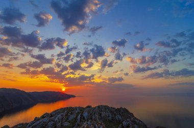 Doğu Sibirya. Baykal Gölü. Aya Körfezi. Pembe, turuncu, sarı sayısız kümülüs bulutu zümrüt ve mavi gökyüzünde yükselen güneş tarafından aydınlatılıyor. Güneşin ışıkları. Sudaki yansımalar.