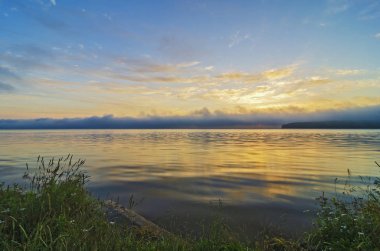 Doğu Sibirya. Baykal bölgesi. Güzel turuncu, sarı, pembe gün doğumu yoğun siste. Sudaki arka plandaki bulutların yansımaları.