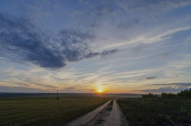 Doğu Sibirya. Baykal bölgesi. Yaz. Bulutlu gökyüzü Güzel turuncu gün batımı. Geniş bir tarlanın kenarındaki köy yolu..