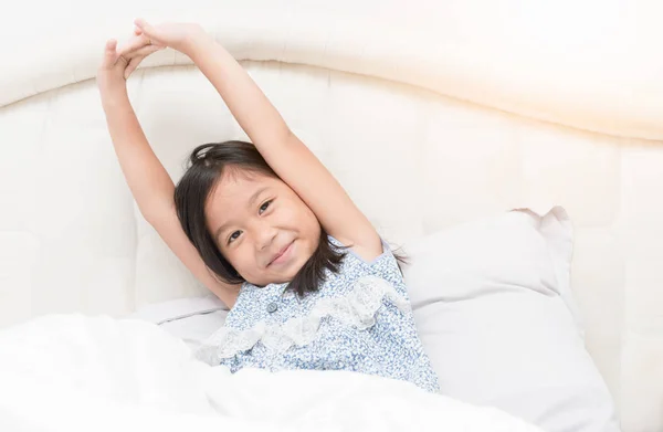 Cute asian girl in pajamas is sitting on  bed stretching. — Stock Photo, Image