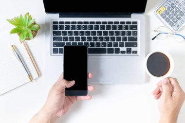 Business man holding smart phone and hot coffee on white office desk table with laptop computer, cup of coffee and vintage bag. Top view with copy space, flat lay.