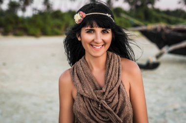 Hippie girl watches and smiles. Beautiful young woman at beach looking at camera. Portrait of attractive brunette girl with long hair. Tanned girl smiling at camera. Joyful girl