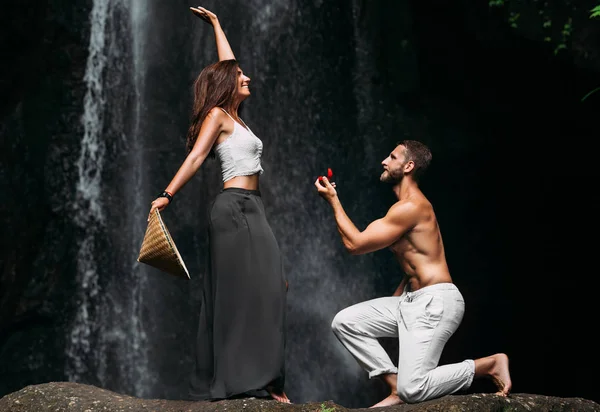 A man proposes to a woman at a waterfall. A guy proposes to a girl in Bali. Offer of a hand in travel. Honeymoon trip. Declaration of love. A couple in love on a waterfall. Happy couple in Bali