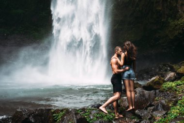A man of athletic build kisses a beautiful girl at the waterfall. Honeymoon trip. Declaration of love. A couple in love on a waterfall. Happy couple in Bali. Beautiful couple travels the world