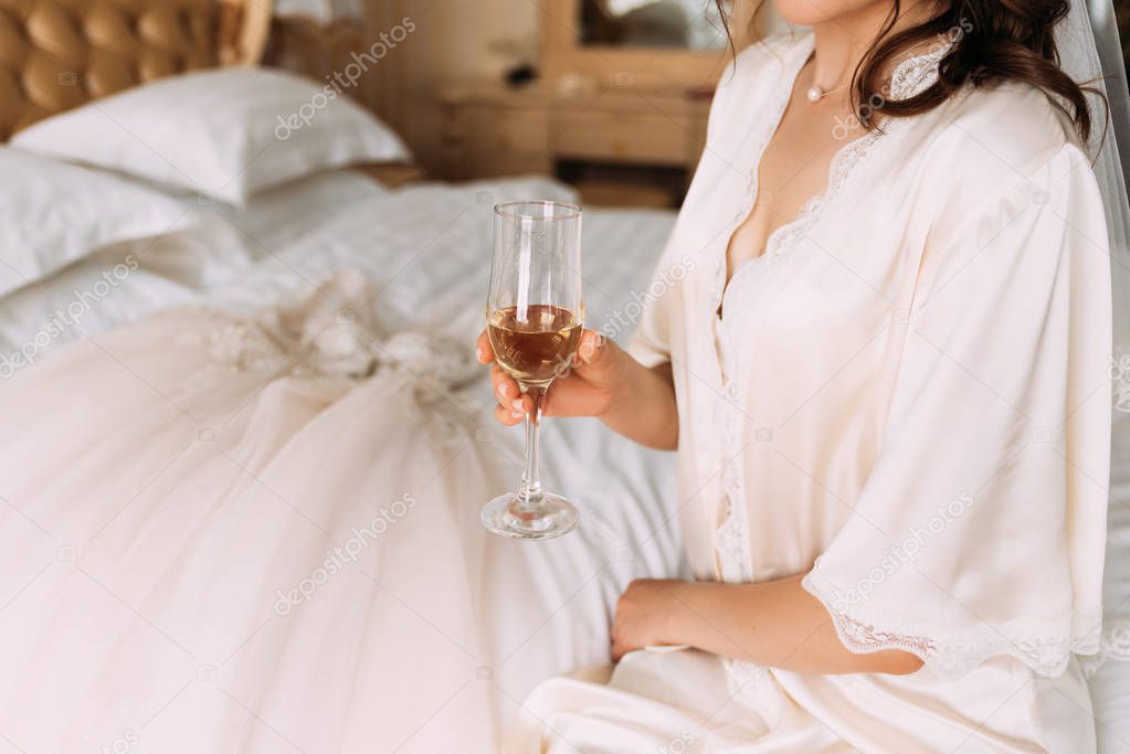 Bride`s morning. Bride drinking champagne in the peignoir. The bride is drinking champagne in a hotel room. Beautiful young woman in white wedding robe holding champagne glass in hand. Bridal morning