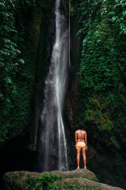 Bali adasındaki bir şelalede sarı şortlu esmer bir adam. Bir adam dünyayı dolaşır. Şelaledeki adam, arkadan görüş. Bali Endonezya 'ya seyahat. Doğanın arka planındaki adam.